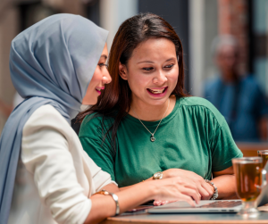 Two Asian women smiling and working on a laptop, one is wearing a head scarf